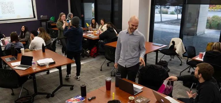 Students talking to each other in a classroom sitting or standing around square table desks.