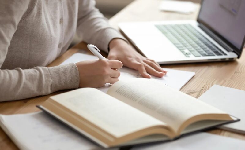 Person writing on a piece of paper with an open book and computer on the desk