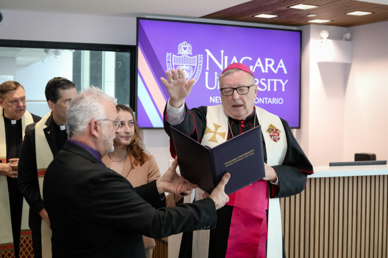 Father blessing while Mike Jeswald is holding a Niagara University hardcover folder