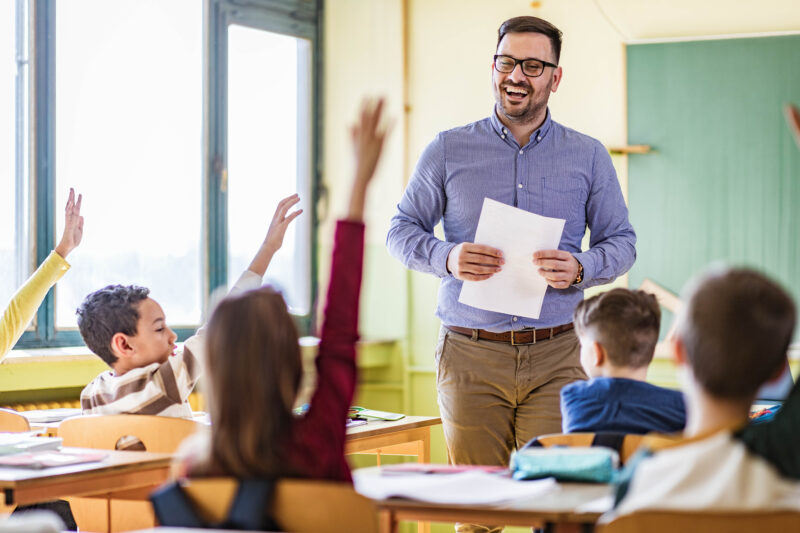 Happy male teacher looking at his students who raised their hands to answer the question on a class at elementary school.