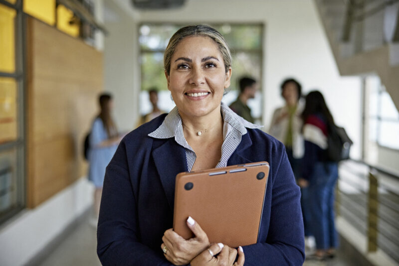 Waist-up view of late 40s woman standing in university hallway, holding laptop, and smiling at camera. Property release attached.